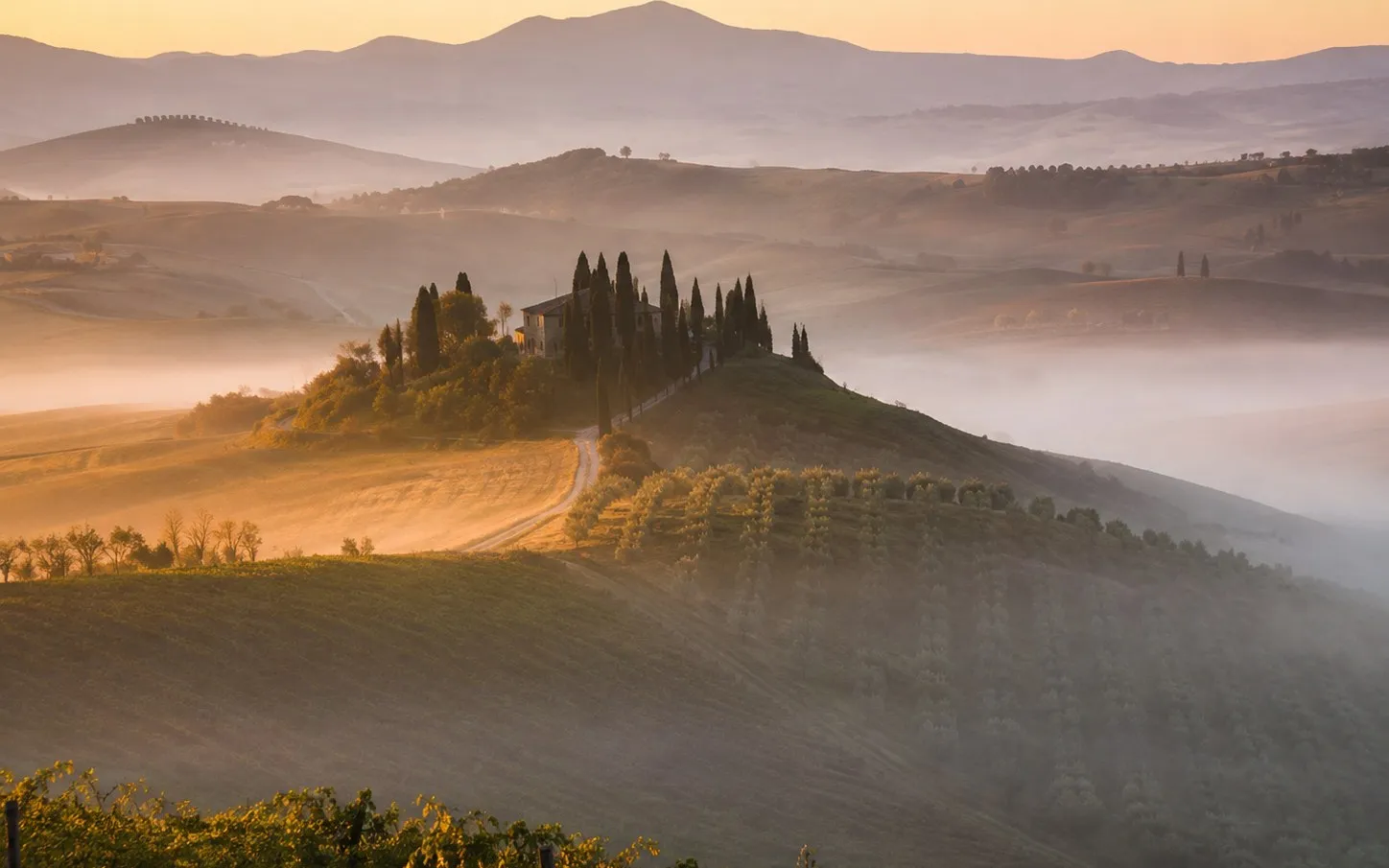 Lone Tuscan farmhouse on a ridge above a misty hollow at sunrise, warm light on the walls, rolling hills behind, wide landscape