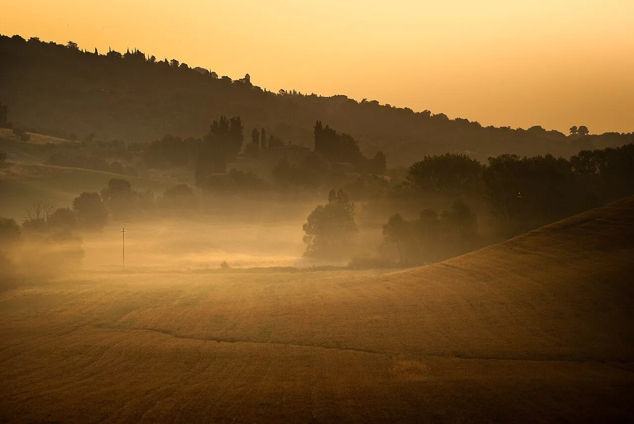 Morning mist over the Val d'Orcia at sunrise with a farmhouse and cypress trees catching the first warm light, wide landscape