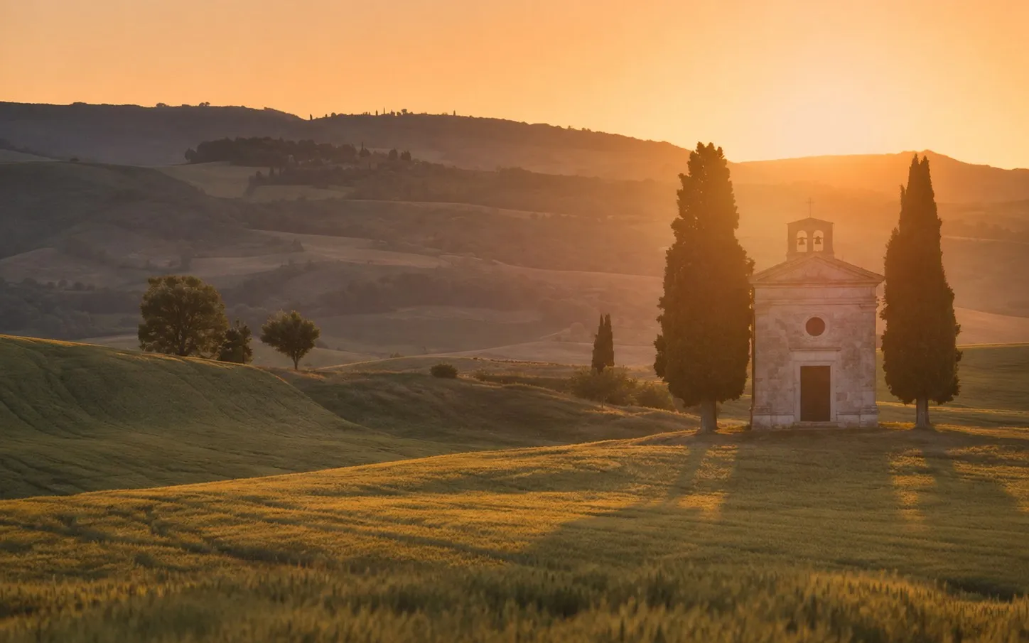 Small stone chapel standing alone flanked by a tall cypress on each side in the Val d'Orcia at golden hour, wide landscape